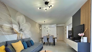 Una sala de estar moderna con un sofá gris, una mesa de centro redonda, un comedor con mesa y sillas, y un televisor montado en la pared.