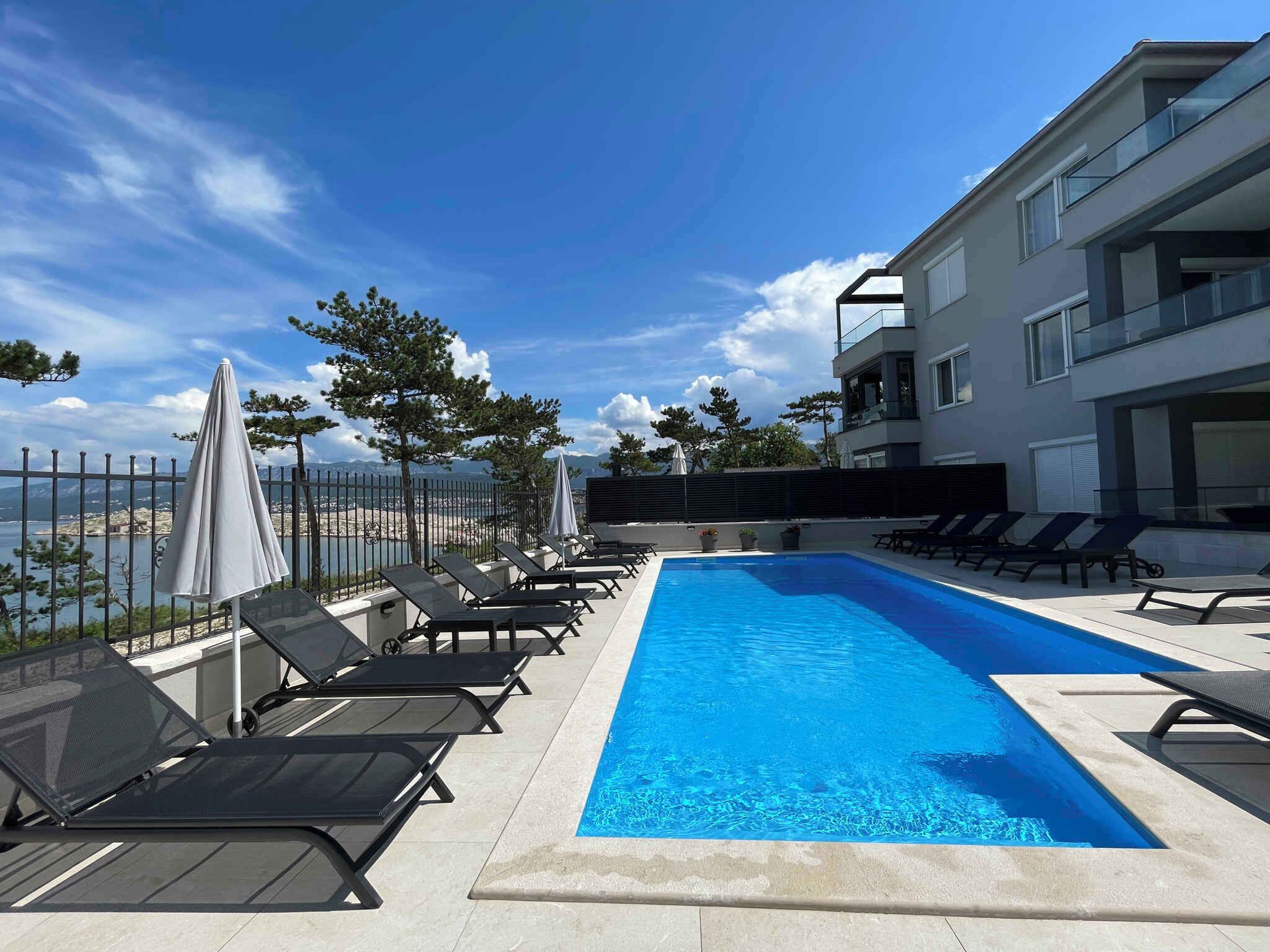 Ein moderner Poolbereich mit Liegestühlen und Sonnenschirmen mit Blick auf ein Gewässer und eine Berglandschaft.