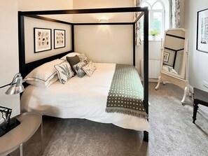 Een hemelbed met een wit en groen beddegoed, meerdere kussens en ingelijste botanische prints aan de muur.