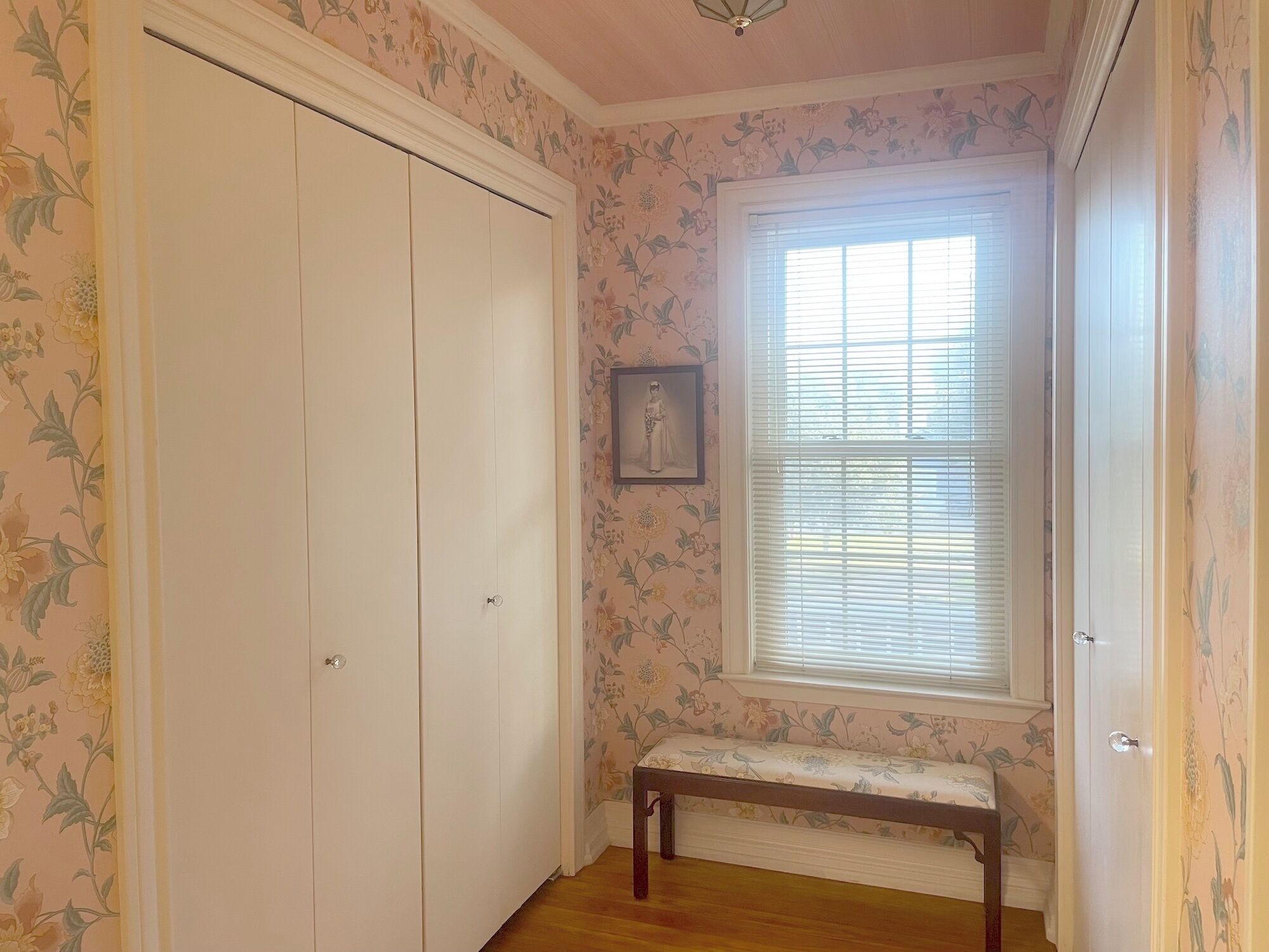 A room with a large white closet, a window with blinds, a bench, and floral wallpaper.