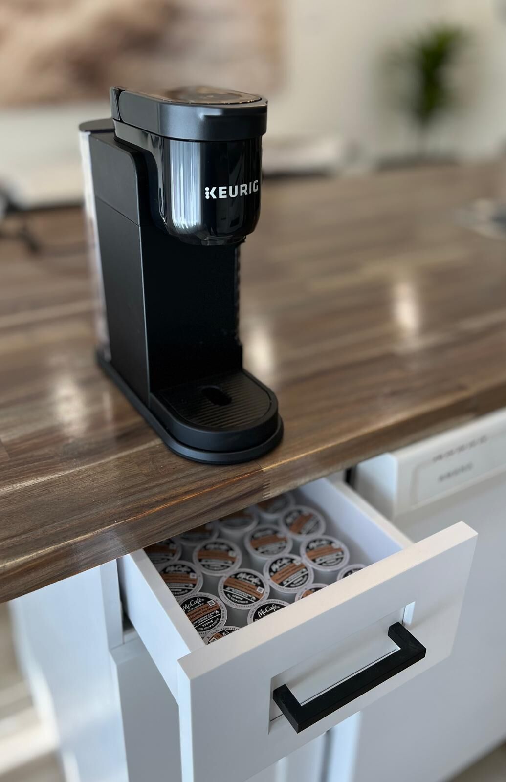 A Keurig coffee machine on a wooden countertop with a drawer below containing coffee pods.