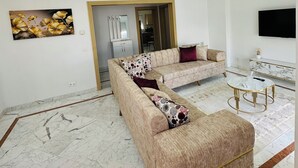 A modern living room with a beige sectional sofa, a round coffee table, and a flat-screen TV mounted on the wall. - Stunning 3 Bedroom Apartment in Khezema, Sousse Just 5 Minutes From the Beach! (Sousse)
