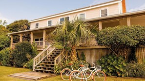 Uma casa de dois andares com escadaria de madeira que dá para uma varanda, cercada por vegetação exuberante e uma bicicleta estacionada na frente.