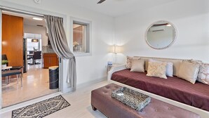 A bedroom with a bed, a bedside table, a lamp, a ceiling fan, a window with blinds, and a door leading to another room.