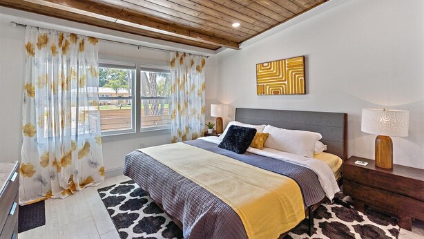 A bedroom with a wooden ceiling, a bed with a grey headboard, two bedside lamps, and a large window with floral curtains.