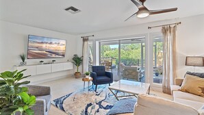 A spacious living room with a large flat-screen TV, a ceiling fan, and a view of the outdoors through large sliding glass doors.