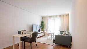 Uma sala de estar moderna com parede de tijolos brancos, piso de madeira, sofá cinza, escrivaninha branca com cadeira, poltrona pendente e uma grande janela com cortinas.