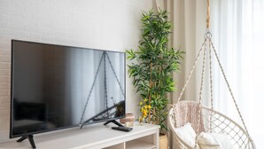 Uma televisão de tela plana sobre um suporte branco, uma cadeira suspensa e uma planta verde alta com flores amarelas.