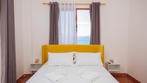 A hotel room with a yellow headboard, white bedding, and a view of the sea through a window with grey curtains. - Villa Gaudium (Orikum)