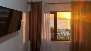 A room with a TV mounted on the wall, a window with curtains, and a view of the sea and buildings at sunset. - Villa Gaudium (Orikum)