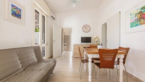 A modern living room with a sofa, dining table, and chairs. There is a TV and a clock on the wall. - 3960 Villa Colle Azzurro by Barbarhouse (Porto Cesareo)