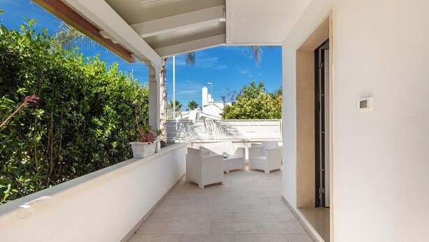 A covered outdoor patio with white wicker furniture, a tiled floor, and a view of a building with a white facade and a blue sky. - 3960 Villa Colle Azzurro by Barbarhouse (Porto Cesareo)