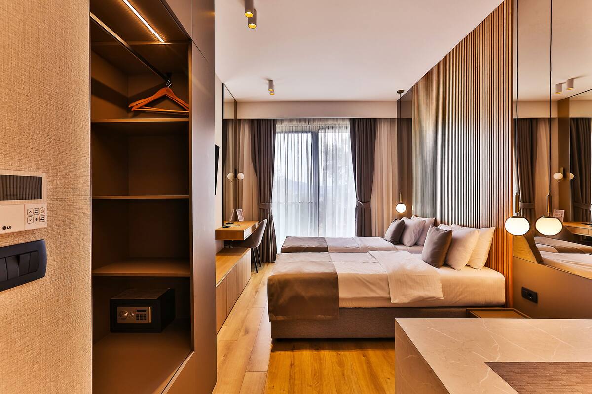 A modern hotel room with a wooden floor, a bed with white linens, a desk with a chair, and a wardrobe.