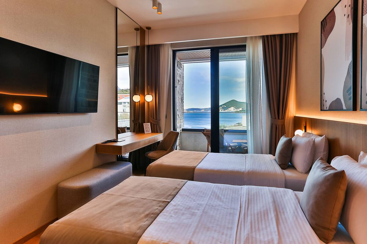 A hotel room with two beds, a desk, a chair, and a TV. There is a large window with a view of the sea and a mountain.