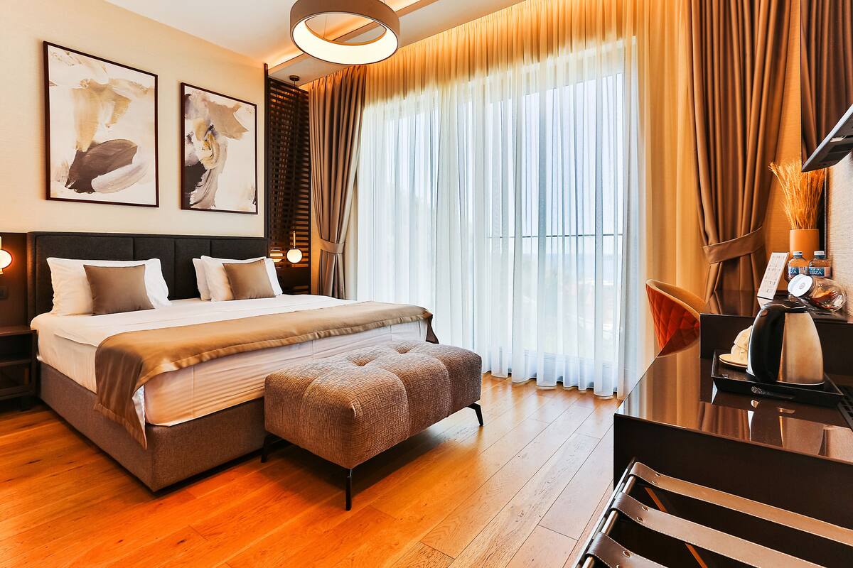 A modern hotel room with a large bed, a bedside table with a lamp, and a view of the city through sheer curtains.