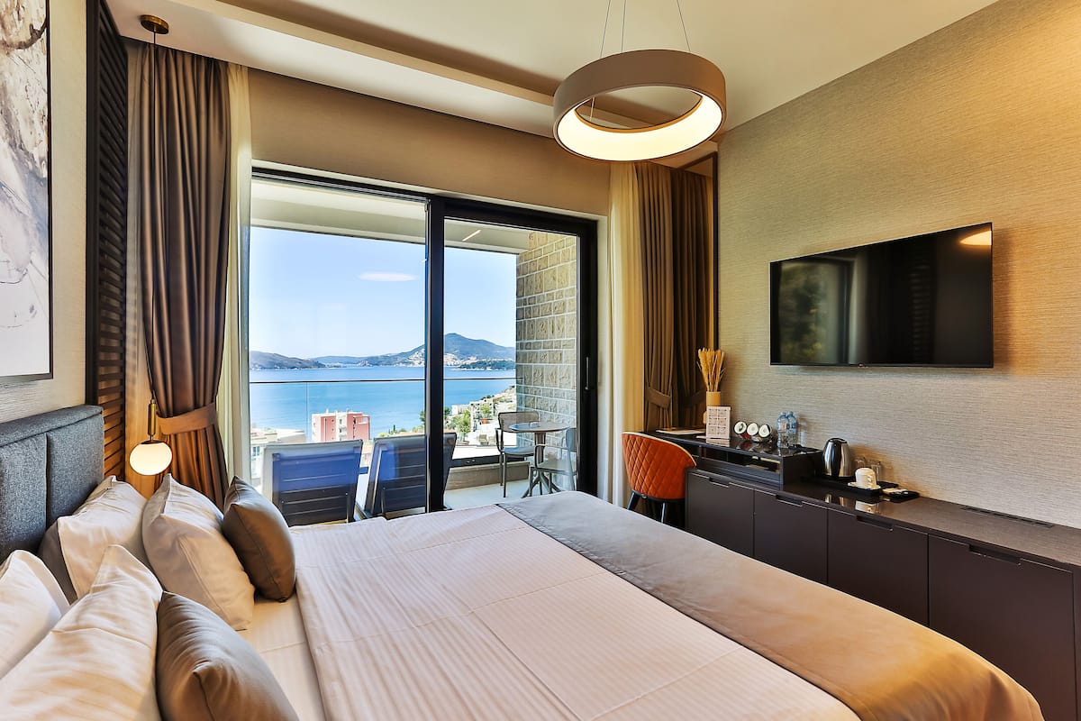 A hotel room with a bed, a desk, a chair, a TV, and a balcony with a view of the sea.