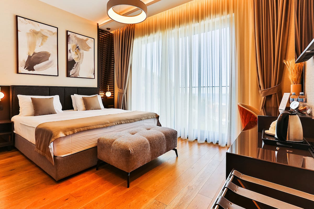 A modern hotel room with a large bed, a bedside table with a lamp, and a view of the city through sheer curtains.