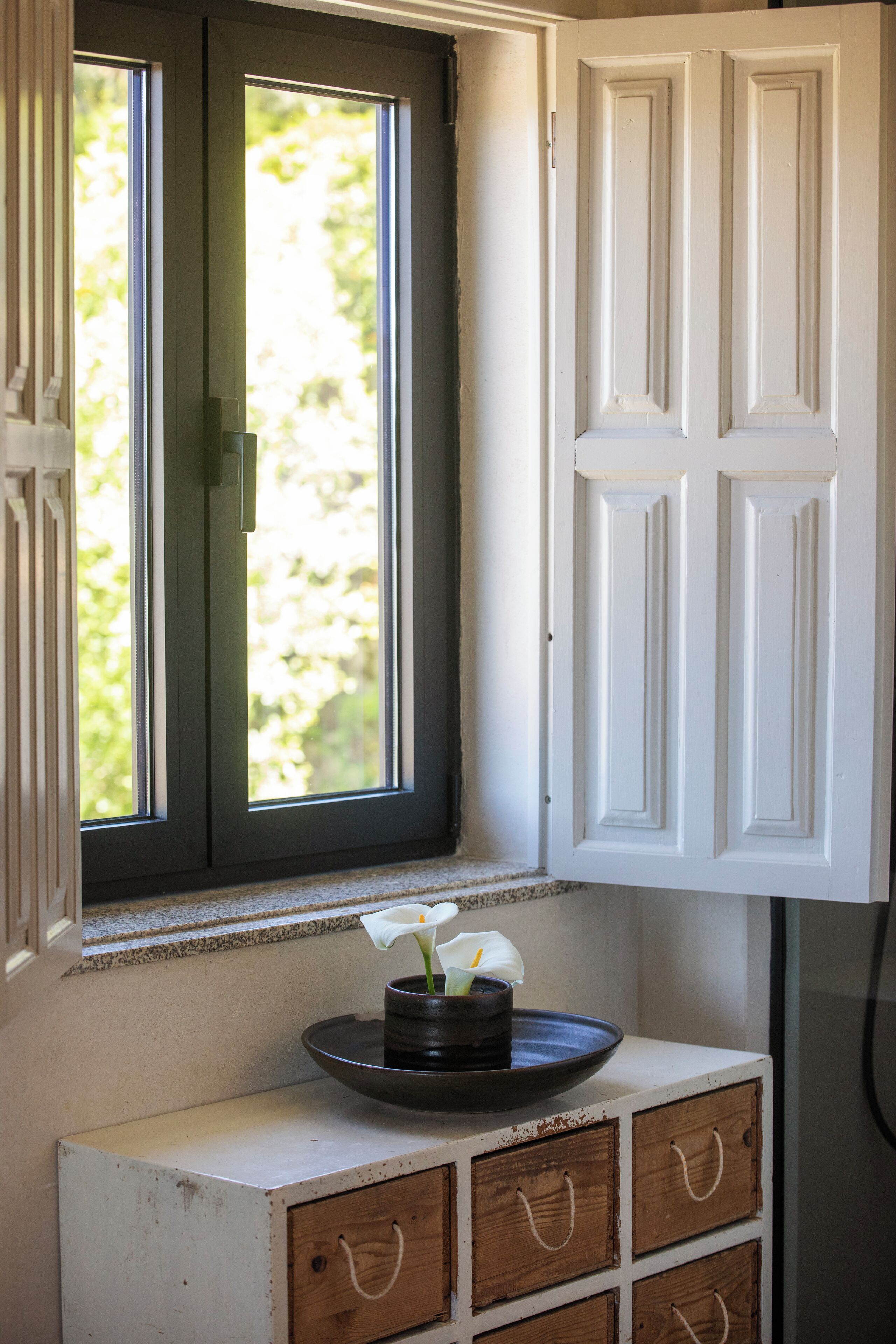 Une fenêtre donnant sur un paysage verdoyant, un meuble blanc avec des tiroirs et une petite table sur laquelle est posé un vase de fleurs.