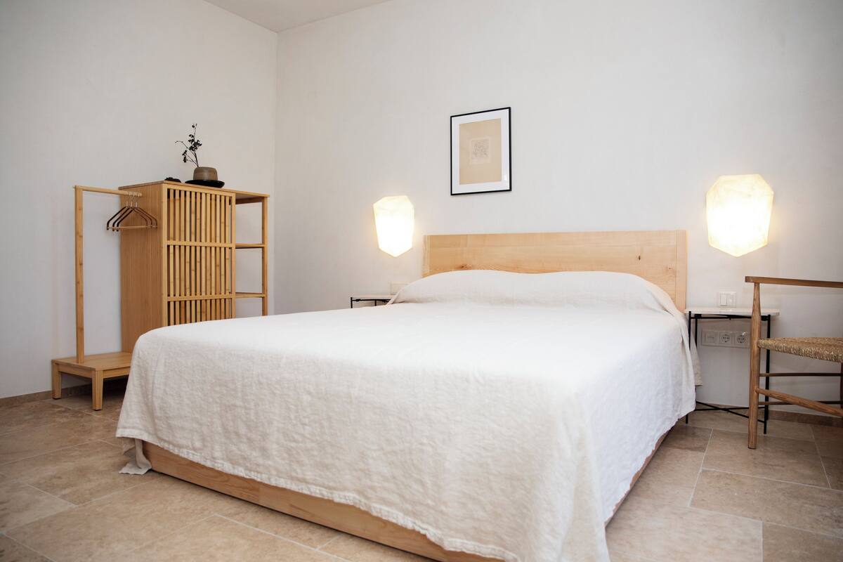 Ein schlicht eingerichtetes Schlafzimmer mit einem hölzernen Bett, einem Nachttisch, einem an der Wand montierten Regal und einem Kleiderhaken.