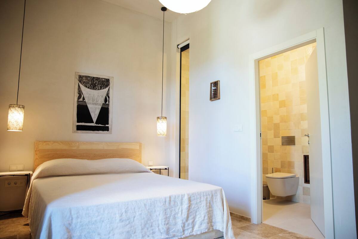 Ein Schlafzimmer mit Bett, Nachttischen, einem an der Wand hängenden Kunstwerk und einem Badezimmer mit Toilette und gefliesten Wänden.