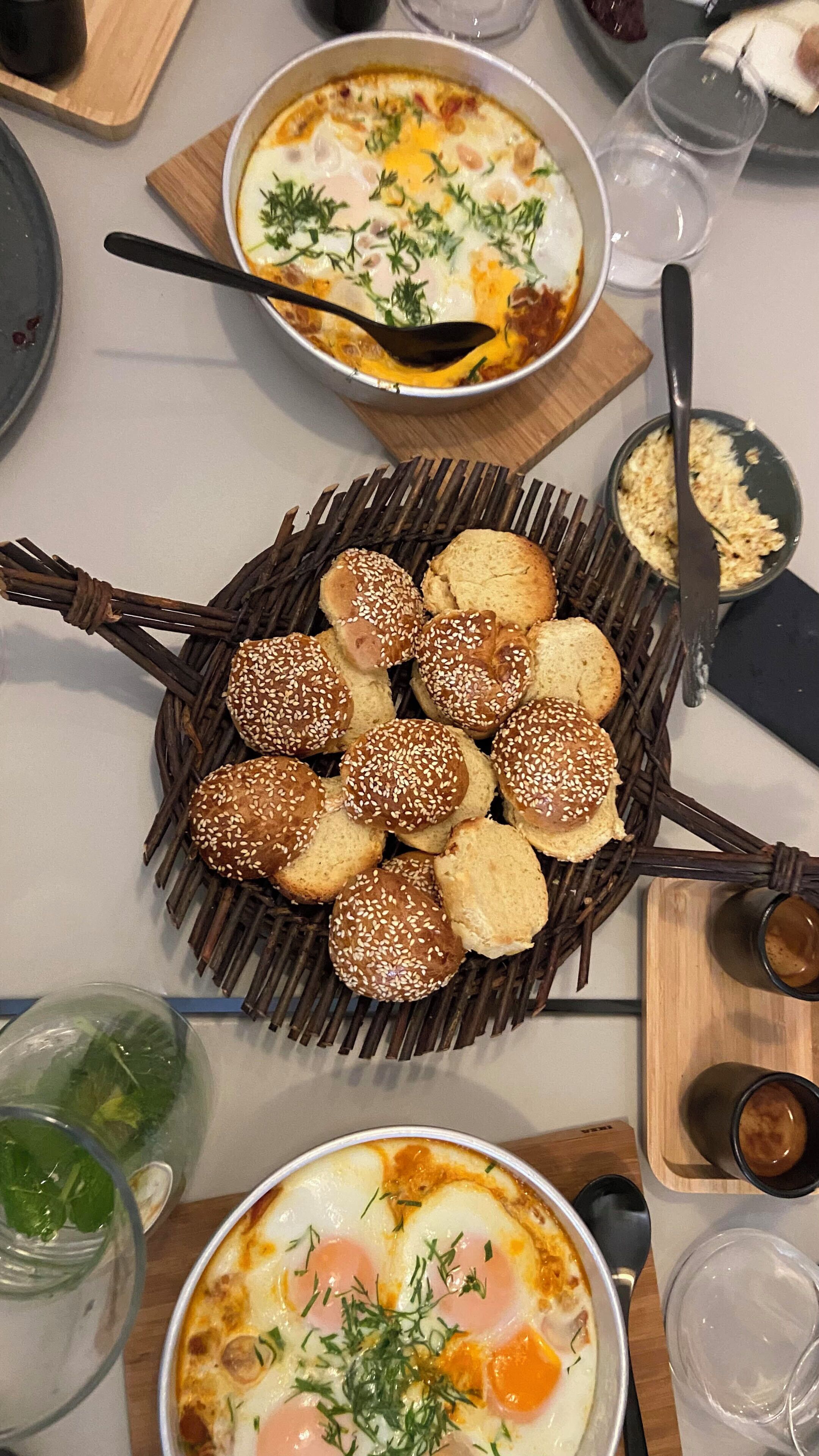 A dining table with various dishes, including a bowl of soup, a plate of eggs, and a basket of sesame seed buns.