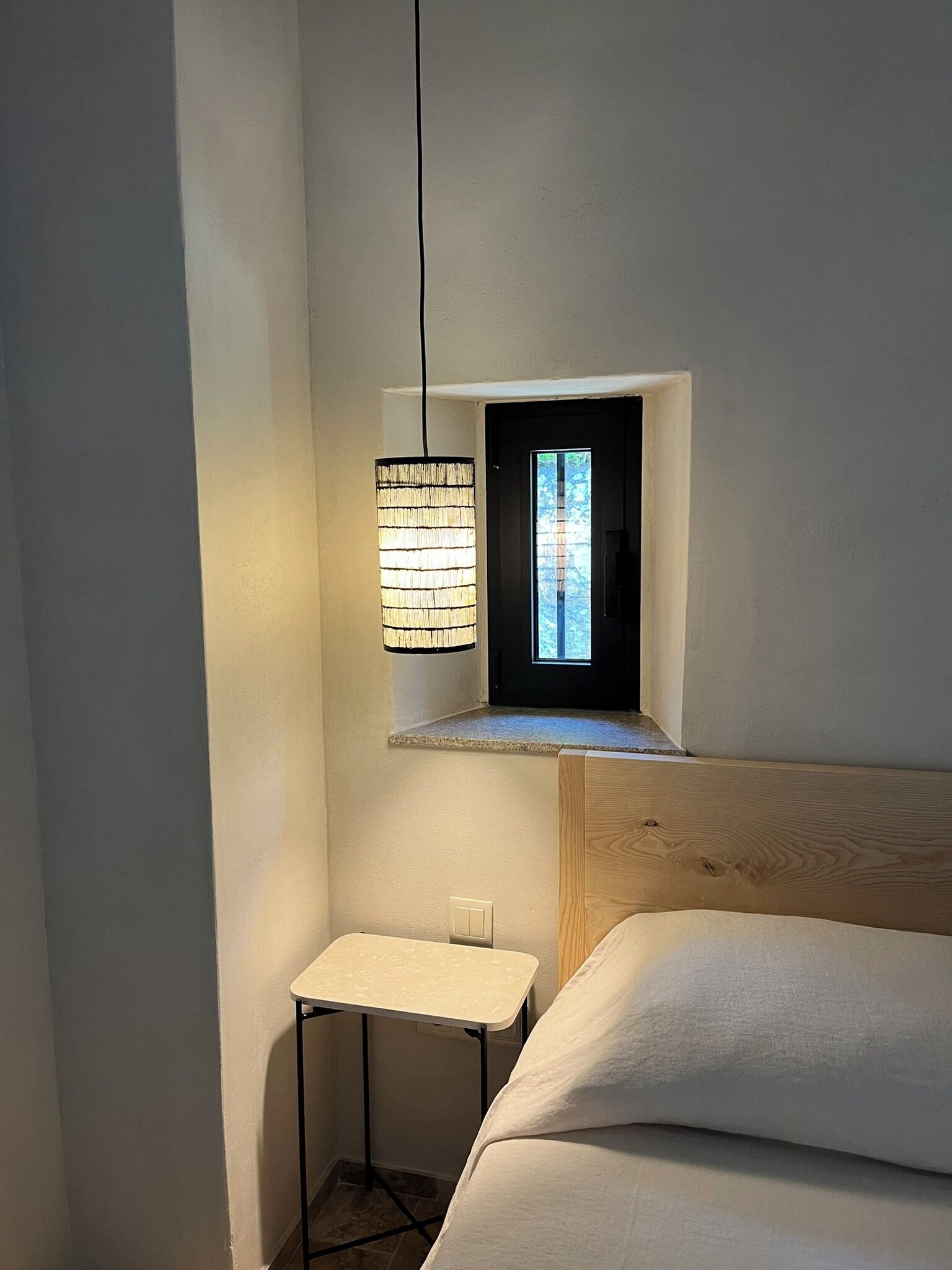 Un dormitorio con una pequeña ventana, una mesita de noche y una lámpara colgante.