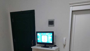 Una habitación con una puerta blanca, una puerta negra, un armario blanco con un televisor encima y un termostato pequeño fijado a la pared.