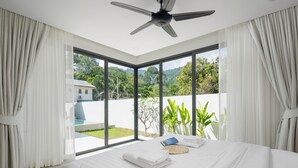 Un dormitorio moderno con una cama grande, un ventilador de techo y vistas a un jardín exuberante y un edificio a través de ventanales de gran altura.