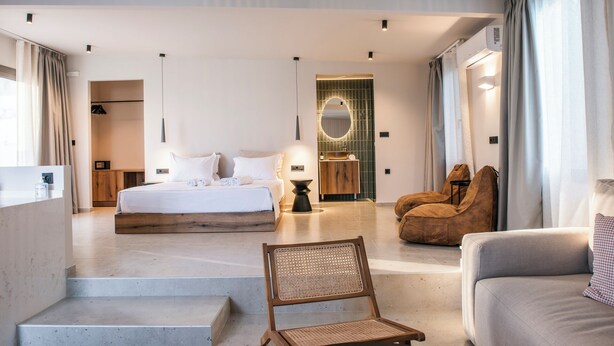 Et moderne hotelværelse med en stor seng, et siddeområde med stol og sofa, et lille bord og et badeværelse med spejl og vask.