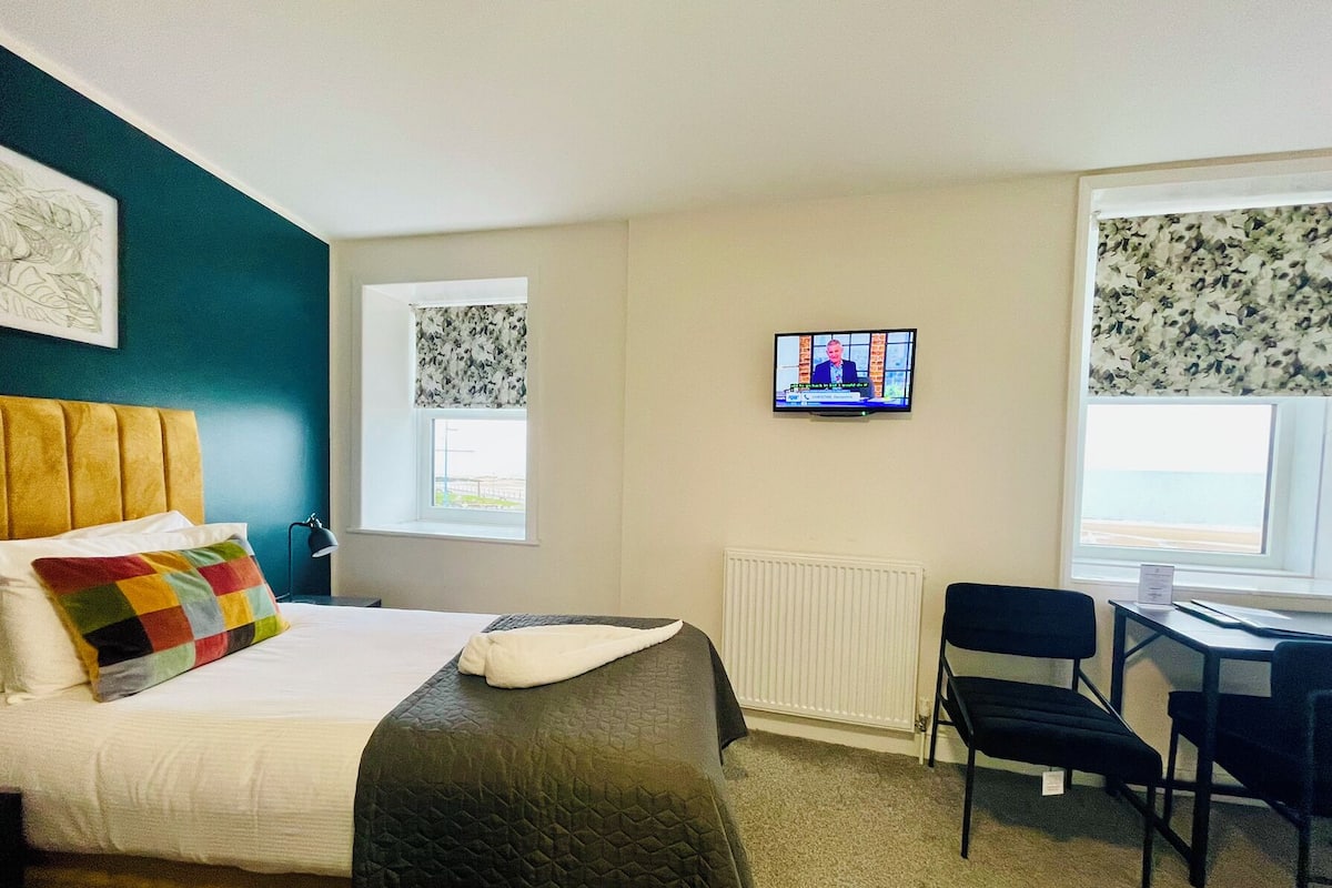 A hotel room with a bed, a desk, a chair, a television, and two windows with floral curtains.