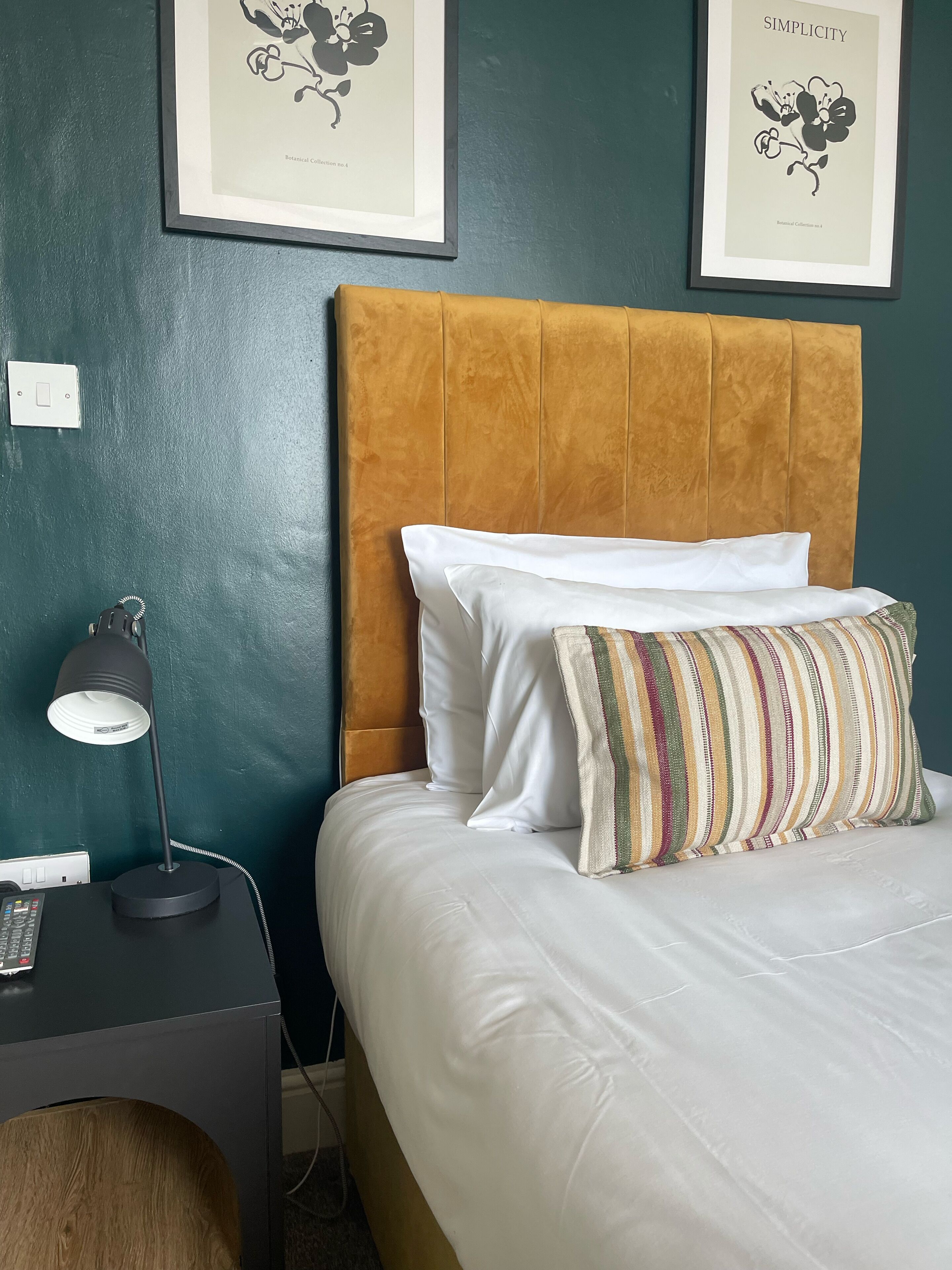 Una cama bien hecha con sábanas blancas y almohadas a rayas, una lámpara de noche e impresión enmarcadas de motivos botánicos en la pared.