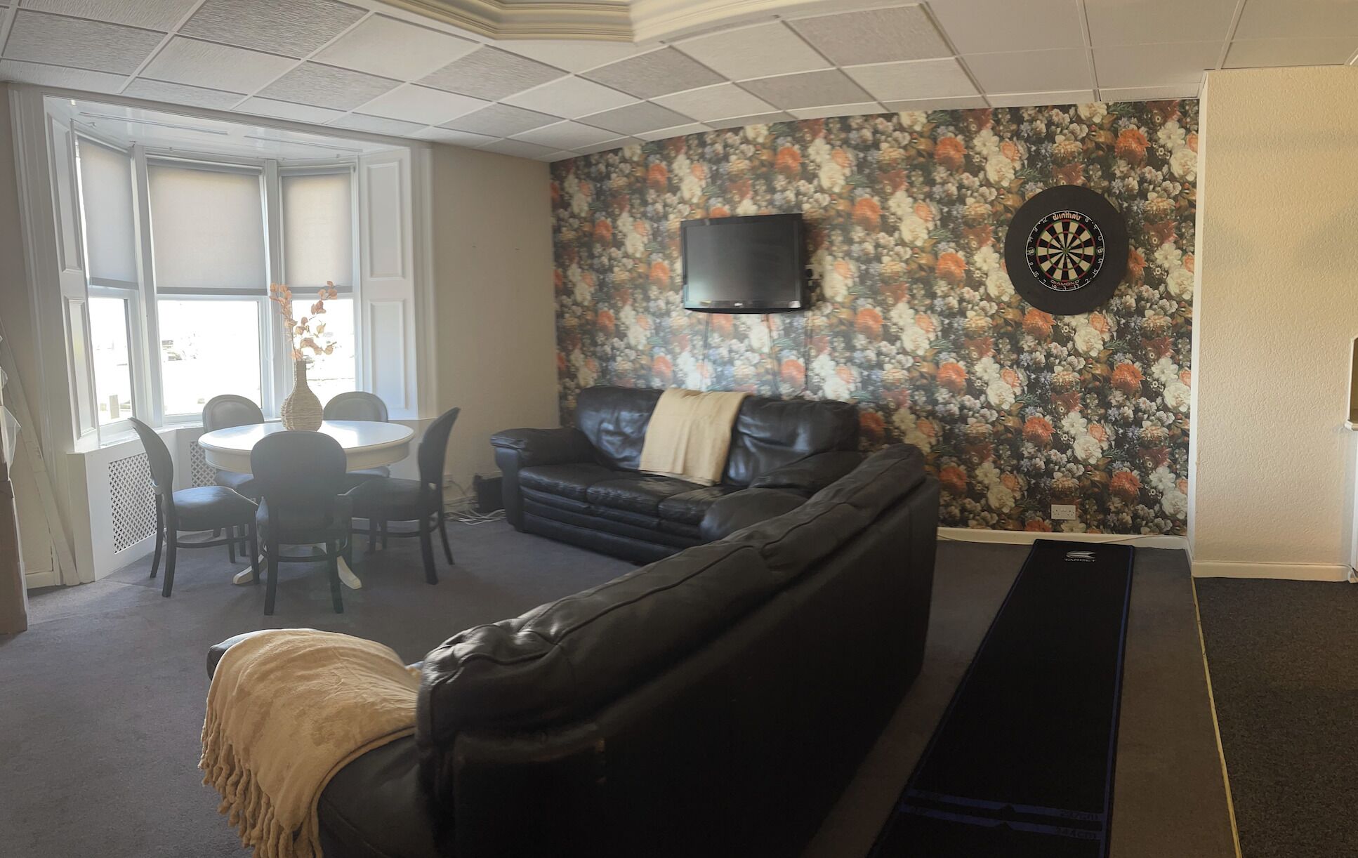 A living room with a floral wallpaper, a black leather sofa, a TV mounted on the wall, and a dartboard.