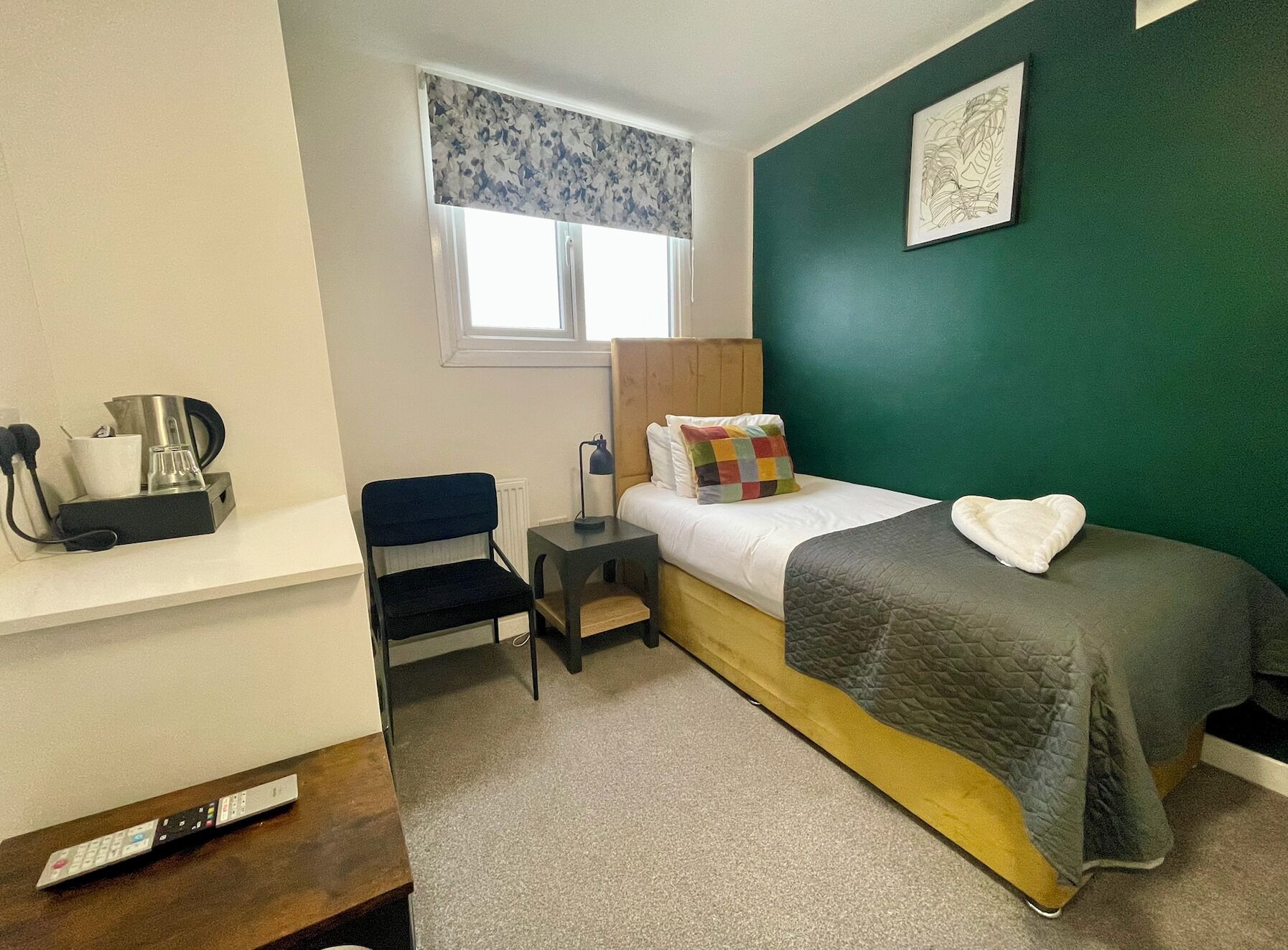 A small, neatly arranged room with a bed, a chair, a desk with a coffee maker, and a window with a floral curtain.