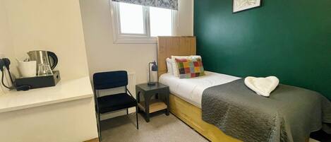 A small, neatly arranged room with a bed, a chair, a desk with a coffee maker, and a window with a floral curtain.