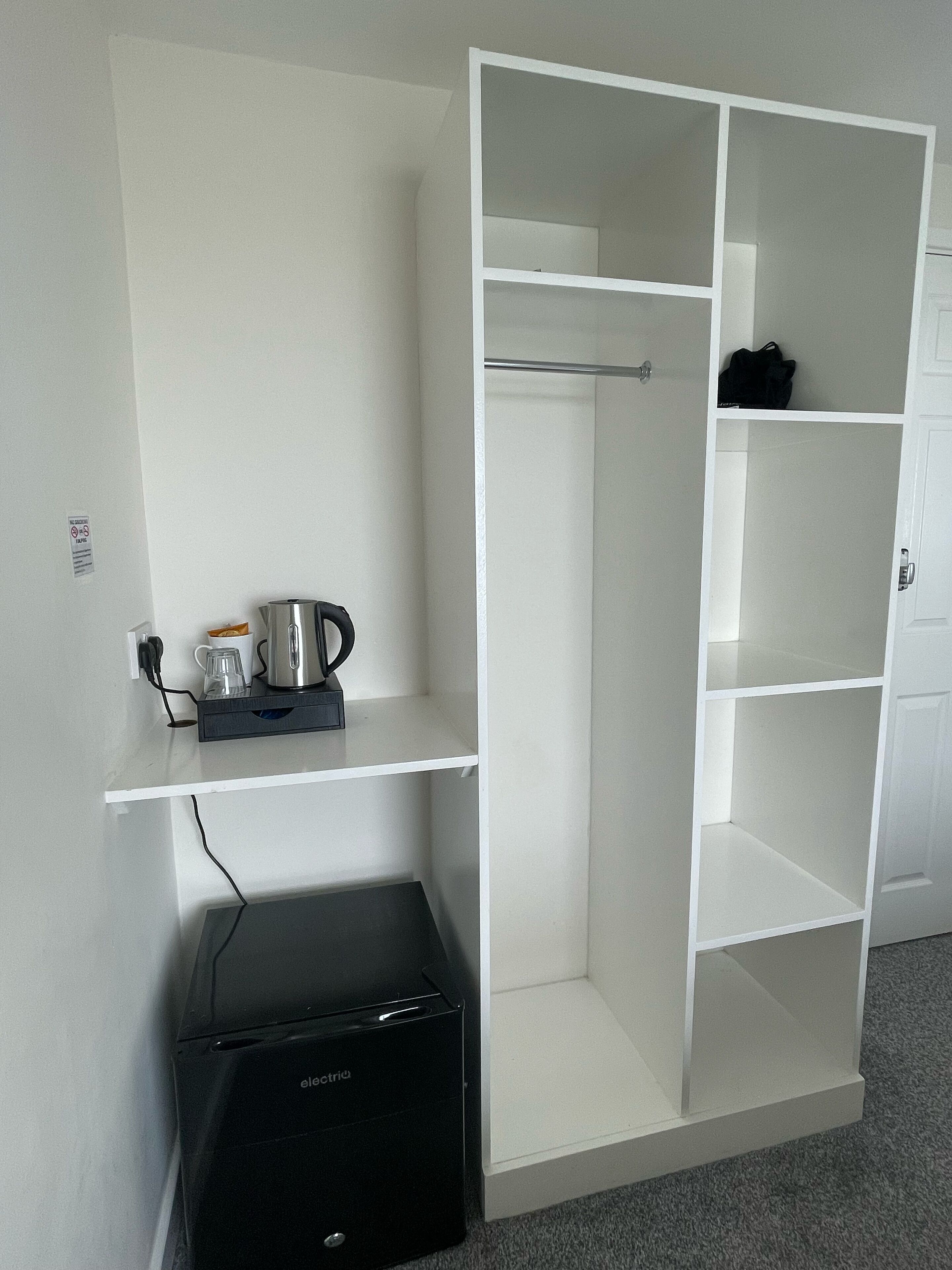 A white wardrobe with multiple shelves and hanging space, a black printer, and a small shelf with a coffee maker and glasses.