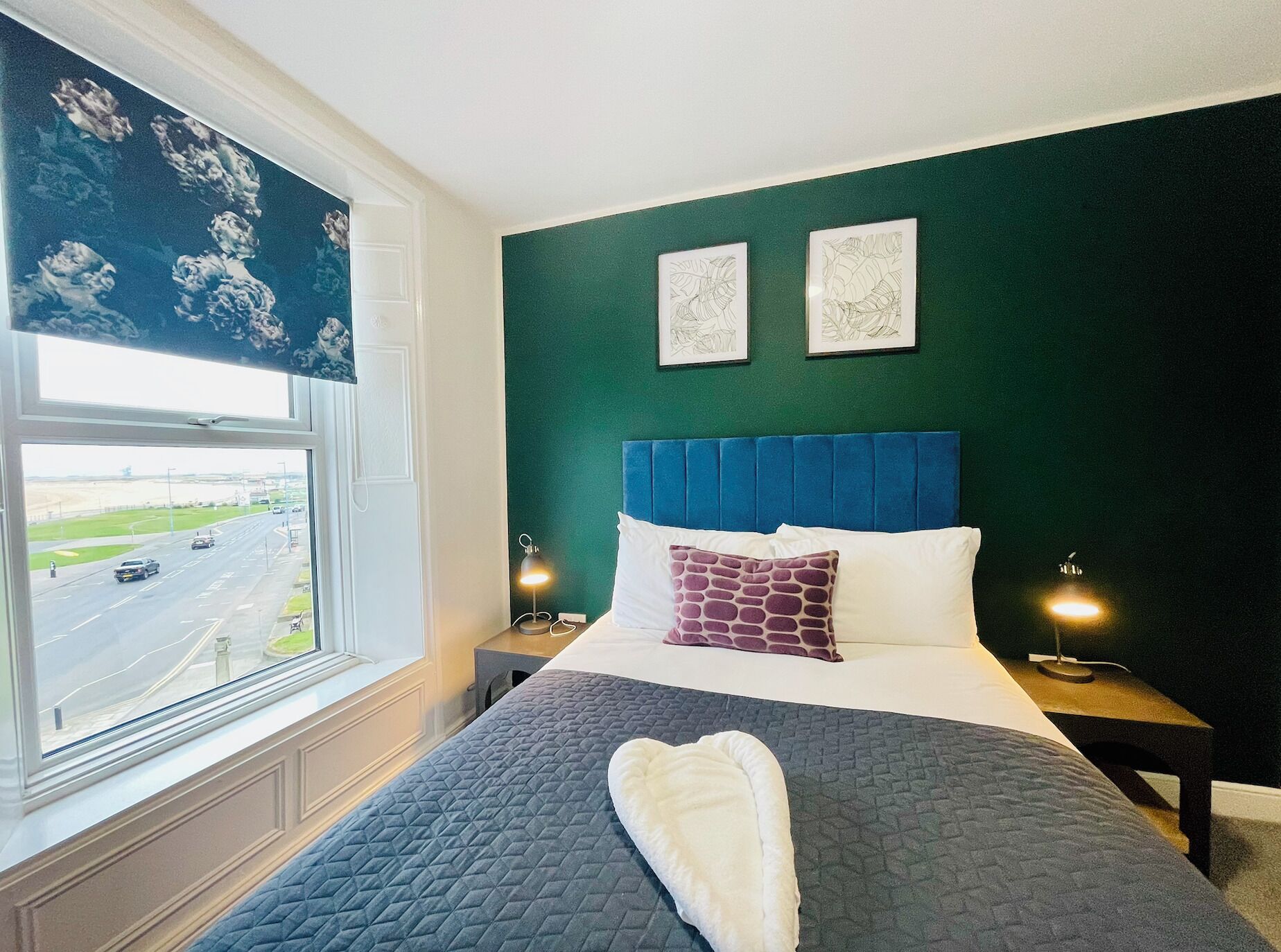 A hotel room with a large bed, a view of a highway through the window, and two framed artworks on the wall.