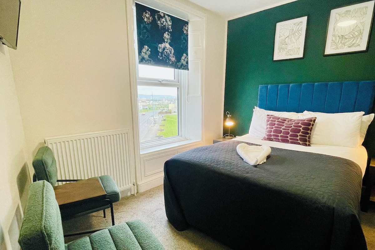 A hotel room with a bed, a chair, a radiator, a window with a view, and two framed pictures on the wall.