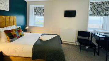 A hotel room with a bed, a desk, a chair, a TV, and two windows with curtains.