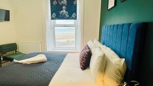 A bed with a blue headboard, a view of the beach through a window, and a green chair. - The Norton (Hartlepool)