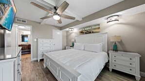A bedroom with a large bed, a ceiling fan, a nightstand with a lamp, and a TV mounted on the wall.
