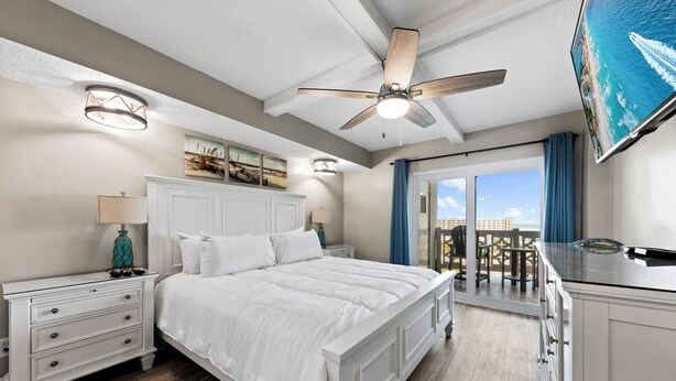 A bedroom with a large bed, a ceiling fan, a TV, and a balcony with a view.