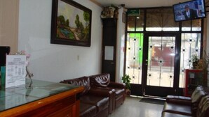 A reception area with a wooden desk, leather sofas, a painting on the wall, a TV on the wall, and a door with a green window. - Hotel Los Angeles  (Tulancingo)