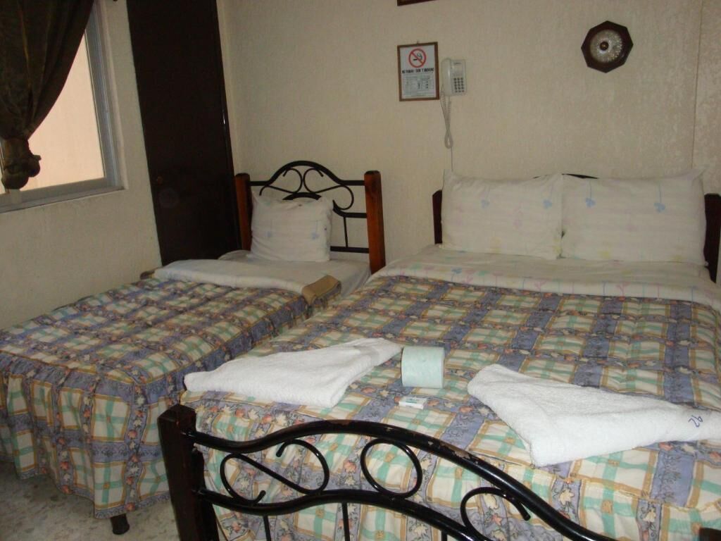 Quarto com duas camas de solteiro, cada uma com colcha estampada e travesseiro branco. Há um telefone na parede e uma porta à esquerda.
