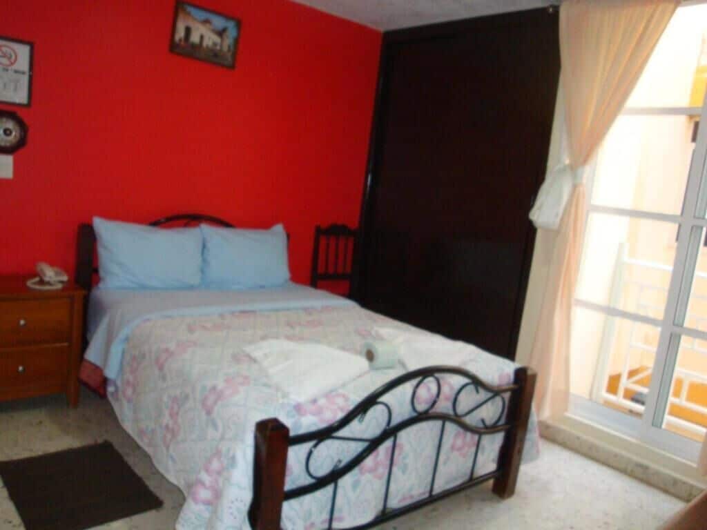 Quarto com cama, mesa de cabeceira, janela com cortinas e parede vermelha.