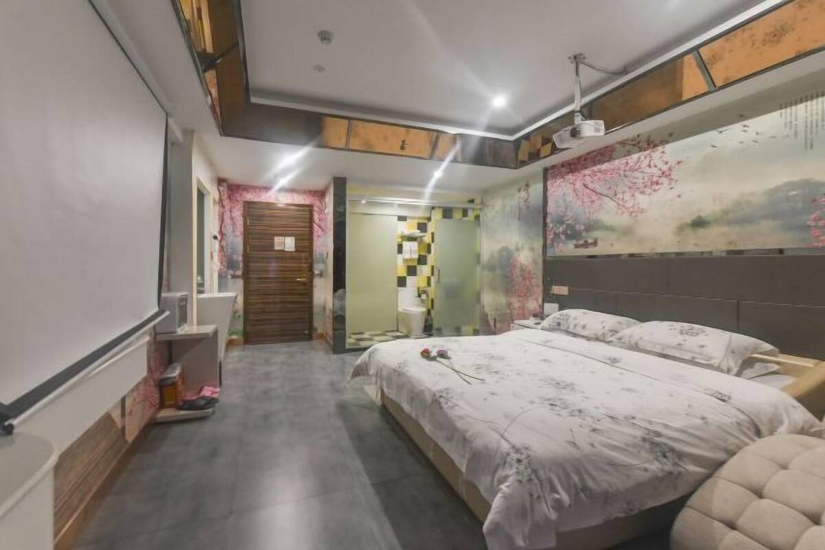 A bedroom with a large bed, a desk, and a chair. There is a door leading to another room.