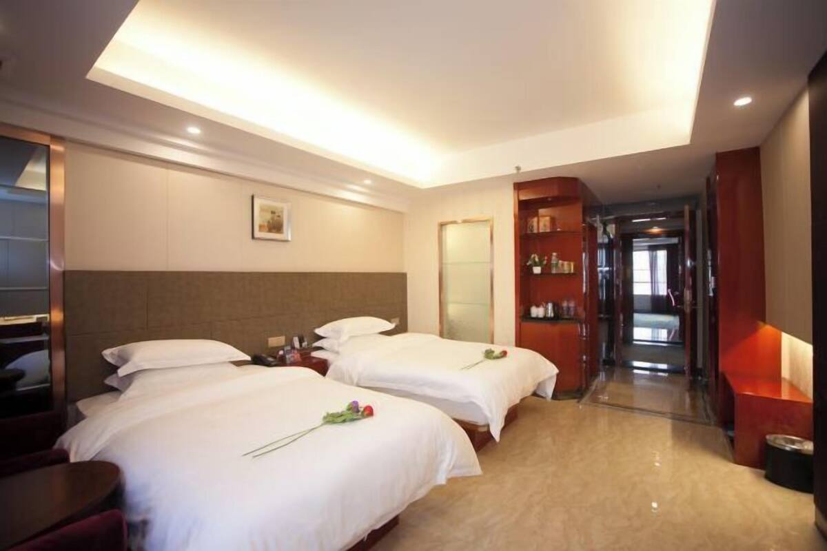 A hotel room with two beds, a desk, a mirror, and a hallway leading to other rooms.
