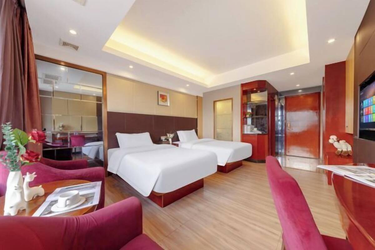 A hotel room with two beds, a sitting area with a red sofa, a wooden floor, a TV, and a small table with a vase and a cup.