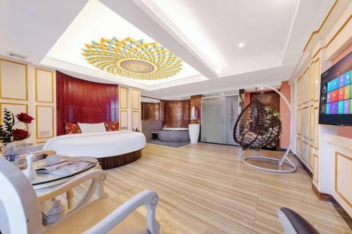 A hotel room with a large bed, a desk with a chair, a TV, a ceiling light fixture, and a wall-mounted TV.