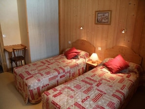 Une chambre avec deux lits simples, chacun doté d’un oreiller rouge et d’une tête de lit en bois. Une petite table avec une chaise se trouve à gauche des lits, et une lampe fixée au mur est placée entre les deux lits.