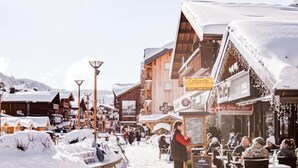 A snowy village scene with wooden buildings, outdoor seating, and a sign for 'Creperie'. - Wifi, Ground floor, parking, tv, ski locker, 15m², Les Gets (Les Gets)